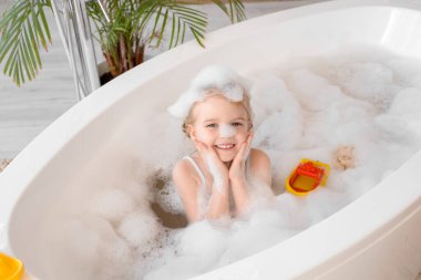 Şirin küçük kız banyo yaparken köpük ve oyuncaklarla banyo yapıyor.