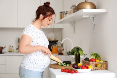 Genç hamile kadın mutfakta salata için taze sebze kesiyor.