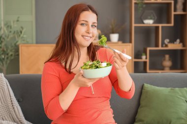 Genç hamile kadın taze sebze salatasıyla evdeki kanepede oturuyor.