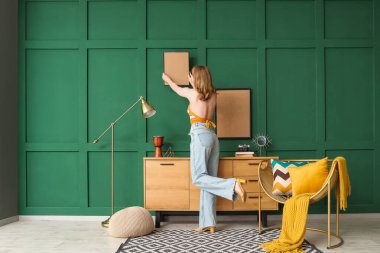 Genç bir kadın yeşil duvara çerçeve asıyor.