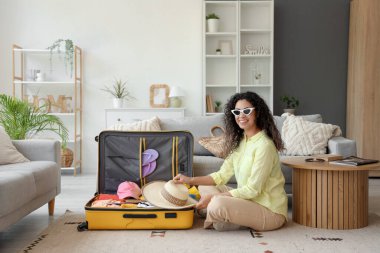 Genç Afro-Amerikalı kadın otel odasında seyahat aksesuarlarıyla bavulunu boşaltıyor.