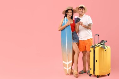 Pembe arka planda bavulu, sörf tahtası ve pasaportu olan mutlu genç çift.
