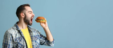 Açık mavi arkaplanda leziz hamburger yiyen aç bir adam.
