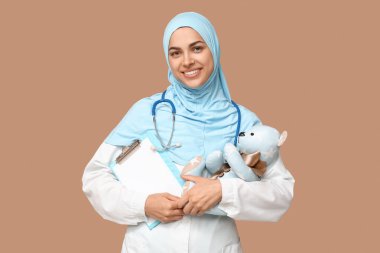 Elinde boş kağıt levha ve bej arka planda oyuncak ayı olan kadın Müslüman doktorun portresi.