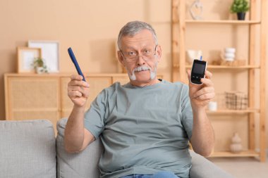 Senior man with glucometer and lancet pen sitting on sofa at home. Diabetes concept clipart