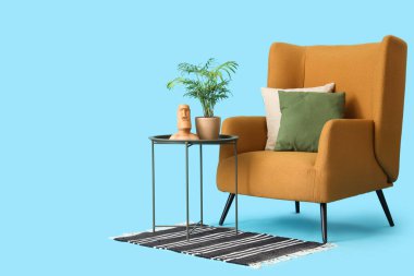 Mavi arka planda bitki ve insan figürlü sehpanın yanındaki yastıklı rahat koltuk.