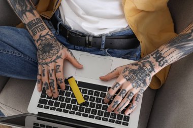 Evdeki koltukta oturan tek kullanımlık elektronik sigara ve dizüstü bilgisayarı olan dövmeli genç bir adam.