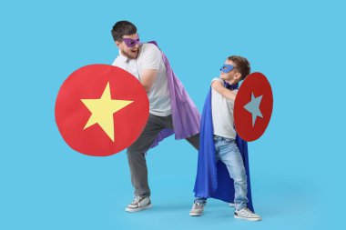 Little boy and his father in superhero costumes with paper shields on blue background clipart