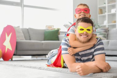 Süper kahraman kostümü giymiş babasıyla evde yerde yatan küçük bir çocuk.