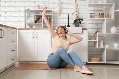 Kulaklıklı genç bir kadın mutfakta yerde oturuyor.
