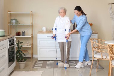 Fizyoterapist yaşlı kadına mutfakta koltuk değnekleriyle yardım ediyor.