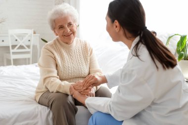 Yatak odasında el ele tutuşan yaşlı bir kadınla fizik terapist.