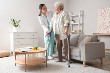 Koltuk değnekleriyle ve fizyoterapistiyle evde yürüyen yaşlı bir kadın.
