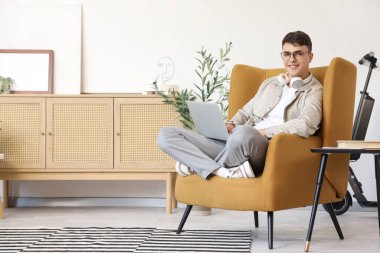 Evdeki koltukta oturan dizüstü bilgisayarlı genç adam.