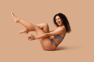 Güzel, genç, mutlu, Afro-Amerikan bir kadın şık iç çamaşırlarıyla bej arka planda oturuyor.