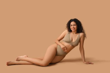 İç çamaşırlı, bej arka planda oturan güzel, mutlu, Afro-Amerikan kadın.