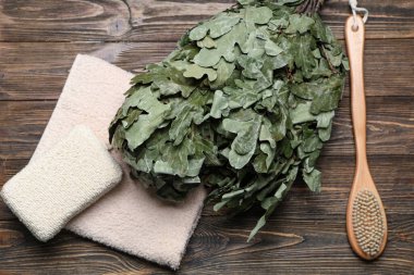 Meşe saunası süpürgesi, masaj fırçası, sünger ve havlu ahşap arka planda.