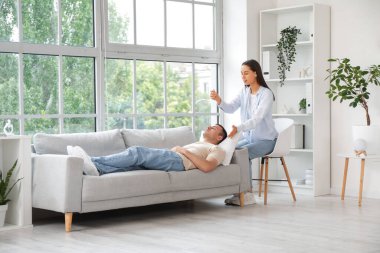 Genç adam psikolog muayenehanesinde hipnoz seansı sırasında kanepede uzanıyor.