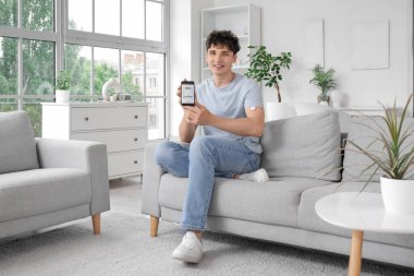 Glikoz sensörü ve cep telefonu olan şeker hastası genç bir adam evdeki kanepede kan şekerini ölçüyor.