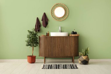 Seramik lavabo, ayna ve ahşap çekmeceler şık bir banyoda yeşil duvarın yanında.