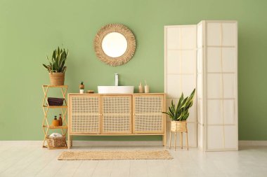 Seramik lavabo, ayna ve çekmeceler şık banyoda yeşil duvarın yanında.