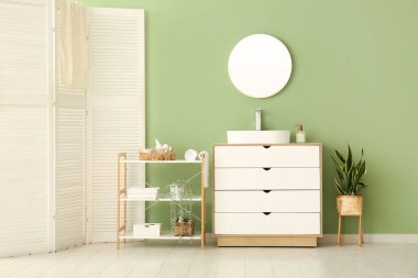 Seramik lavabo, raf ünitesi ve şık banyoda yeşil duvarın yanındaki beyaz çekmeceler.