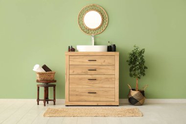 Seramik lavabo, sandalye, ayna ve yeşil duvarın yanındaki ahşap çekmeceler şık banyoda.