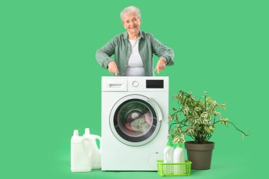 Çamaşır makinesini işaret eden ve yeşil arka planda çamaşır yıkayan mutlu yaşlı kadın.