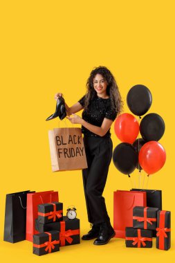 Alışveriş torbaları, hediye kutuları, balonlar ve sarı arka planda çalar saati olan güzel bir Afro-Amerikalı kadın. Kara Cuma indirimi