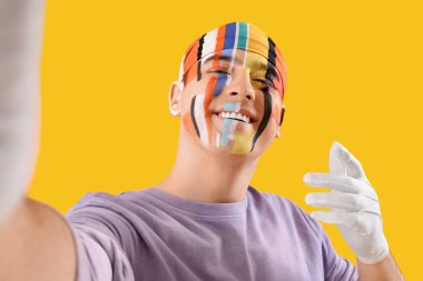 Yüzünde çizgiler olan bandanalı genç adam sarı arka planda selfie çekiyor, yakın plan.