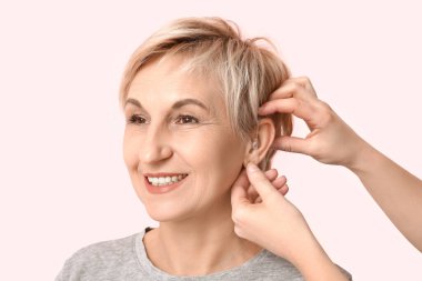 Genç bir kadın pembe arka planda olgun bir kadının kulağına işitme cihazı takıyor.