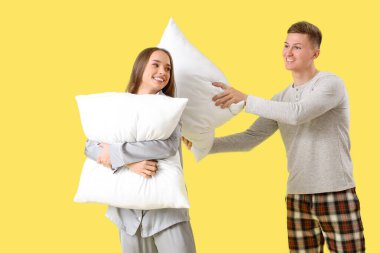 Karısı pijamalı genç adam sarı arka planda yastıklarla savaşıyor.