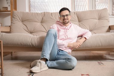 Gözlüklü yakışıklı genç adam evdeki kanepenin yanında oturuyor.