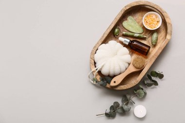 Ahşap tepsi, banyo süngeri, okaliptüs dalı, bir şişe serum, guasha, yüz masajı aracı, mum ve beyaz arka planda deniz tuzu.