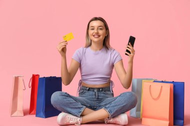 Kredi kartı, cep telefonu ve alışveriş çantaları olan genç bir kadın pembe arka planda oturuyor.
