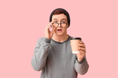 Şapkalı genç adam, pembe arka planda gözlüklü ve bir fincan kahveli.
