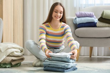 Giysileri yığılmış genç bir kadın evde yerde oturuyor.