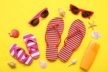 Şık parmak arası terlikleri, güneş gözlüğü, güneş kremi ve sarı arka planda deniz kabukları olan kompozisyon
