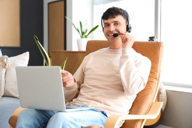 Kulaklığı ve dizüstü bilgisayarı olan genç adam evdeki koltukta konuşuyor.