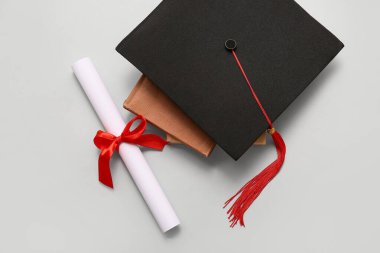 Kırmızı kurdeleli diploma, beyaz arka planda kitap ve mezuniyet şapkası.