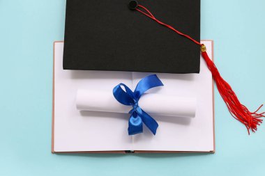 Mavi kurdeleli diploma, açık kitap ve açık mavi arka planda mezuniyet şapkası.
