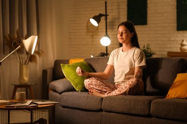 Pijamalı genç bir kadın geceleri oturma odasında rahat bir kanepede meditasyon yapıyor.