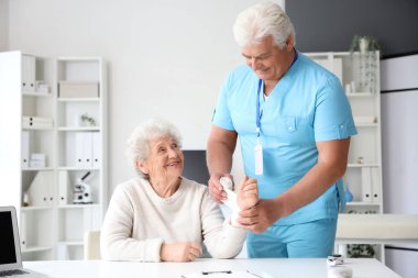 Klinikte erkek doktor yaşlı kadının bileğini bandajlıyor.