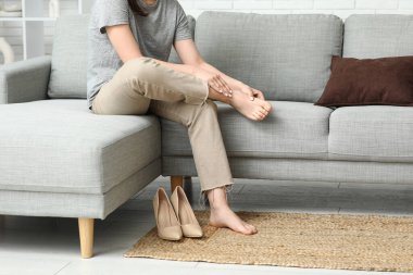 Evdeki gri koltukta topuklu ayakkabı giydikten sonra eklem ağrısı çeken genç bir kadın.