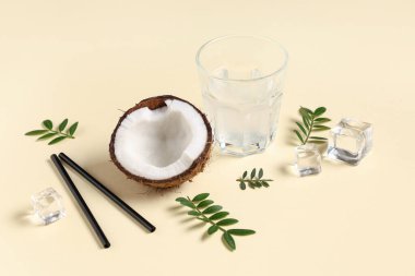 Taze hindistan cevizinin yarısı, bir bardak su, buz küpleri ve sarı arka planda yapraklar.