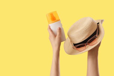 Hasır şapkalı, güneş gözlüklü ve sarı arka planda güneş kremi olan kadın elleri.