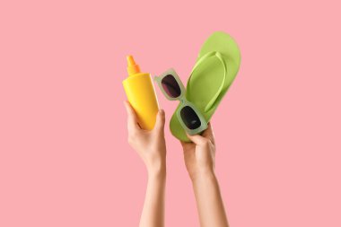 Parmak arası terlik, güneş kremi ve pembe arka planda güneş gözlüğü olan kadın elleri.