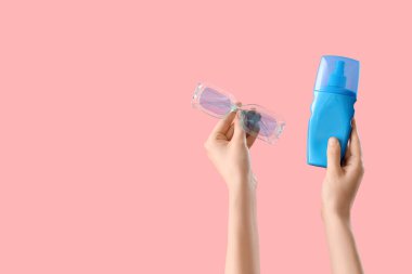 Güneş kremi sürmüş kadın elleri ve pembe arka planda güneş gözlükleri.