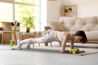 Güzel bir genç kadın oturma odasında yoga minderinde antrenman yapıyor.