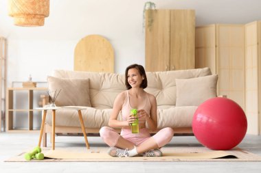 Oturma odasında yoga minderinde spor şişesi olan güzel bir kadın.